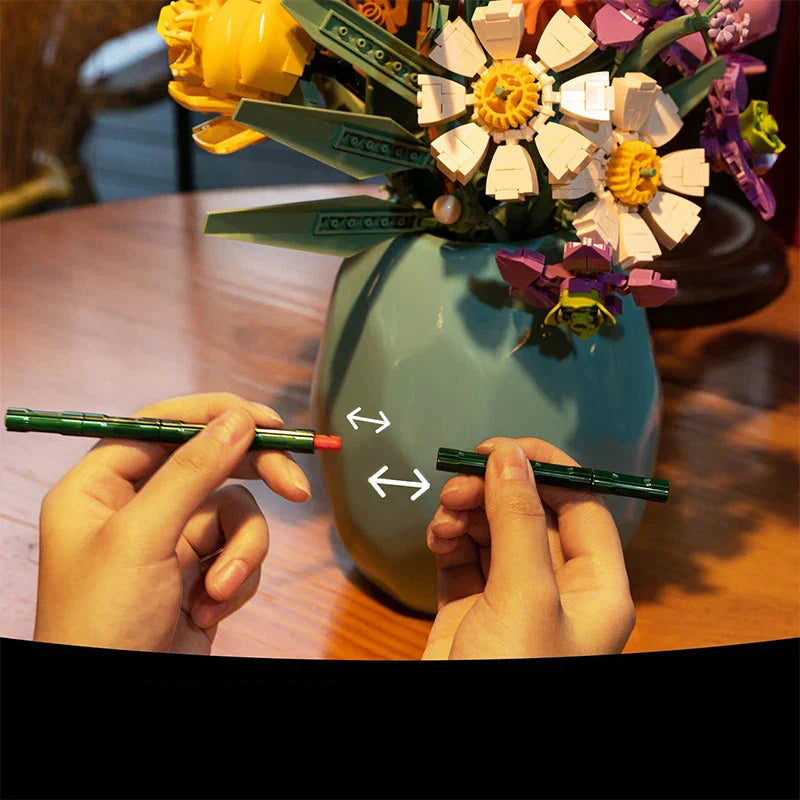 Two hands holding pens with a LEGO flower arrangement in the background.