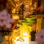 Miniature Japanese street scene with a green waterfall, lanterns, and cherry trees glowing inside a lit book nook