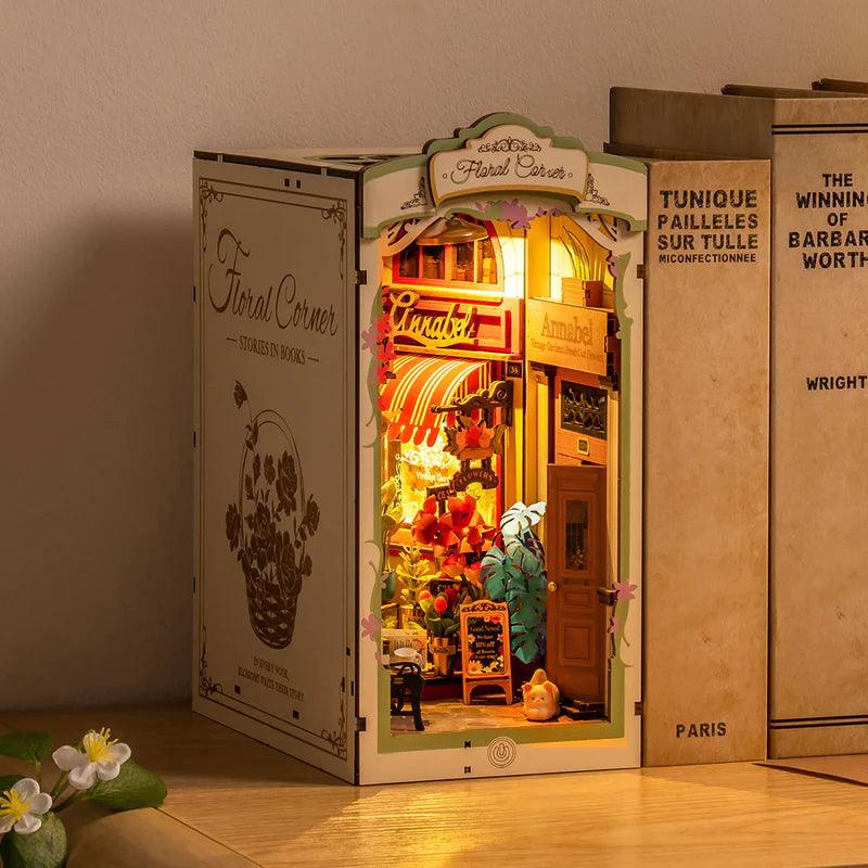 Miniature flower shop book nook slotting between real books as a bookshelf insert, a cozy LED lit gift for book lovers