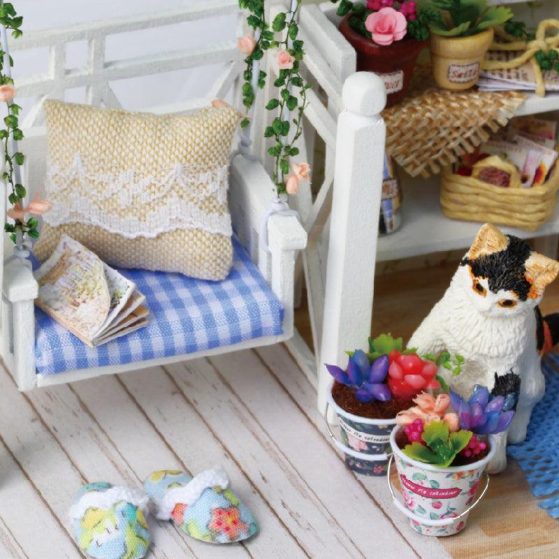 Little porch swing with a lace pillow beside a black and white kitten figure and potted succulents in a cozy dollhouse nook