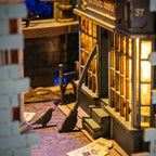 Lit miniature magic shop storefront with a cobbled alley, glowing windows and a witch broom inside the wooden book nook