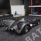 Model of a Batmobile on a dark surface with a framed picture and other items in the background.