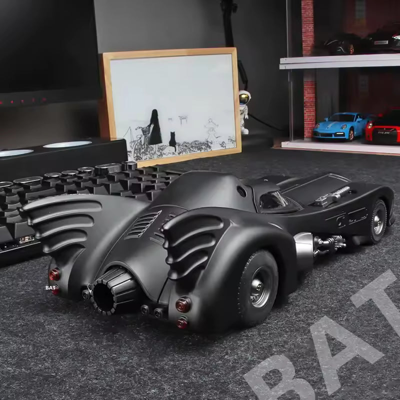 Model of a Batmobile on a dark surface with a framed picture and other items in the background.