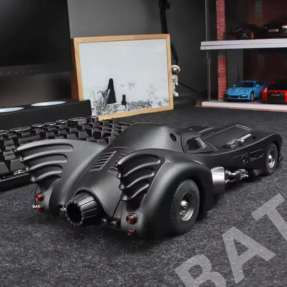 Model of a Batmobile on a dark surface with a framed picture and other items in the background.